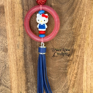Puede incluir: Un amuleto Hello Kitty rosa y rojo con un borla azul. El amuleto está unido a una cuerda blanca y tiene un anillo de purpurina rojo alrededor.