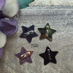 May include: Four star-shaped metal charms with a ring attached to each. The charms are in different finishes: gold, silver, rose gold, and black.