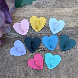 May include: Assortment of ten heart-shaped hooks in various colors, including pink, yellow, blue, green, and black. Each hook has a small ring for hanging items.