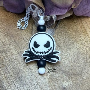 May include: A black and white Jack Skellington-themed charm with a white chain and beads. The charm features a smiling face and a bat-shaped bow tie. The charm is attached to a clear plastic piece.