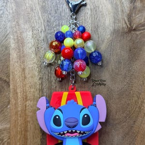 Puede incluir: Un llavero con un personaje de dibujos animados azul, un lazo rojo y un grupo de cuentas de colores. El llavero tiene un cierre plateado y un fondo de madera.