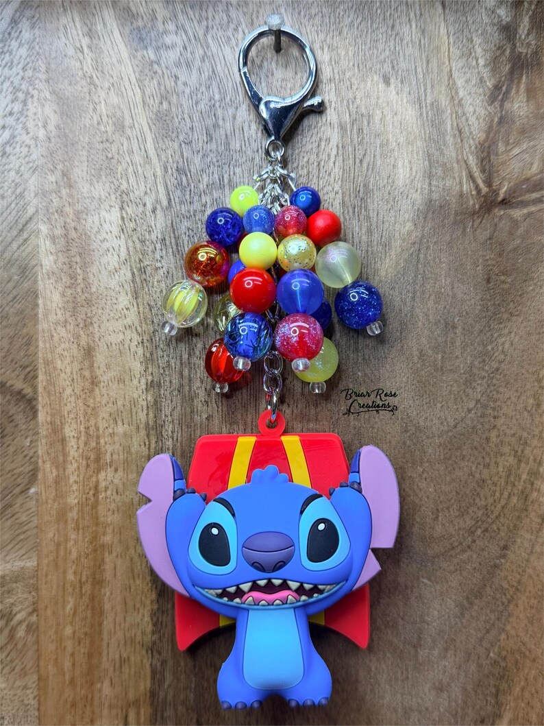 Puede incluir: Un llavero con un personaje de dibujos animados azul, un lazo rojo y un grupo de cuentas de colores. El llavero tiene un cierre plateado y un fondo de madera.