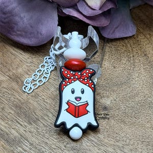 Op de afbeelding: Een witte spookvormige bedel met een rode strik en een boek, op een heldere plastic basis. De bedel heeft een witte ketting en een wit en rood kralenaccent. De geest leest een rood boek. De bedel ligt op een houten oppervlak.