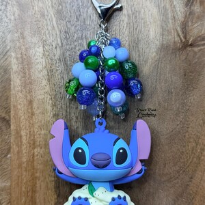 Puede incluir: Un llavero de Stitch azul con un grupo de cuentas azules, verdes y brillantes. El llavero presenta un cierre plateado y el texto "Briar Rose Creations". La figura de Stitch es azul con orejas rosas y una base blanca.