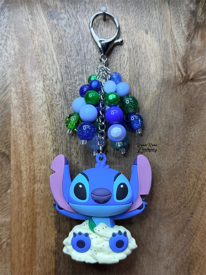 Puede incluir: Un llavero de Stitch azul con un grupo de cuentas azules, verdes y brillantes. El llavero presenta un cierre plateado y el texto "Briar Rose Creations". La figura de Stitch es azul con orejas rosas y una base blanca.