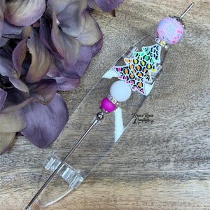 以下が含まれることがあります： ヒョウ柄のデザインが施された、装飾用のクリスマスツリーペン。ペンには、ピンクの花柄ビーズ、白いビーズ、マゼンタ色のビーズが付いています。ペンは透明なアクリルベースにあります。「Briar Rose Creations」の文字が見えます。