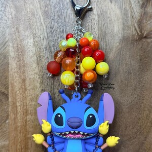 Puede incluir: Un llavero de Stitch azul con un grupo de cuentas coloridas. El llavero presenta un personaje de dibujos animados con ojos grandes, orejas moradas y detalles amarillos. Las cuentas son en tonos rojos, naranjas, amarillos y transparentes.