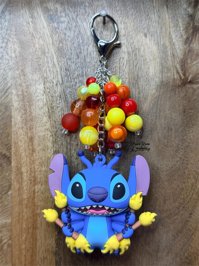 Puede incluir: Un llavero de Stitch azul con un grupo de cuentas coloridas. El llavero presenta un personaje de dibujos animados con ojos grandes, orejas moradas y detalles amarillos. Las cuentas son en tonos rojos, naranjas, amarillos y transparentes.