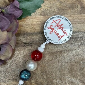 May include: A white Admin Squad badge reel with red script and a beaded accent. The beads are red, white, and blue. The badge reel has the words "Warm, Compassionate, Caring, Loving, Kind, Loyal, Reliable, Fun, Dedicated" around the edge.