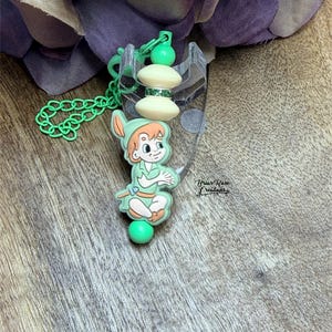 May include: A green and cream-colored keychain featuring a cartoon character. The keychain has a green chain, a green ball, and two cream-colored beads with a silver band. The character is wearing a green outfit and a hat.