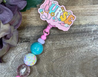 May include: A pink nurse badge reel with the word "NURSE" in colorful letters, a stethoscope, and a syringe. The badge reel has a beaded chain with blue, purple, and clear beads. The background is a wooden surface.