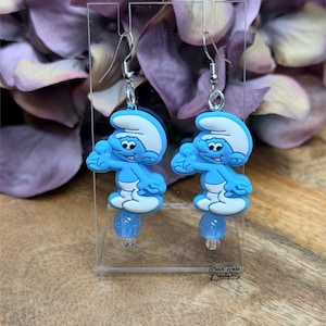 Puede incluir: Un par de pendientes colgantes con personajes de los Pitufos azules con sombreros y detalles blancos. Cada pendiente tiene una cuenta de purpurina azul y ganchos para las orejas plateados. Los pendientes se exhiben en un soporte acrílico transparente.