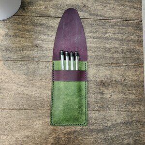 May include: A handmade leather pen case in green and purple, holding four silver pens. The case has a green body, a purple top flap, and a dark purple band. The case is stitched with black thread.