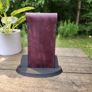 May include: A burgundy leather case with gray stitching, standing upright on a black, rounded base. The case has a smooth texture and a folded-over top. The background includes a plant in a white pot and a wooden table.