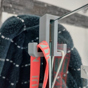 May include: A grey 3D printed hook mounted to a glass shower door. A red and white striped strap is hanging from the hook.