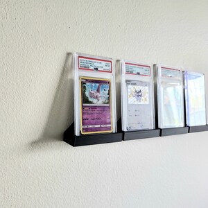Puede incluir: Cuatro cartas de Pok&eacute;mon clasificadas se muestran en un soporte de pared negro. Las cartas est&aacute;n encerradas en soportes de pl&aacute;stico transparente con una etiqueta blanca en la parte superior. Las cartas son todas diferentes, pero todas presentan personajes de Pok&eacute;mon. Las cartas est&aacute;n etiquetadas con el texto "2017 PSA 10 BLACK STAR MEWTWO GOLD STAR 1ST EDITION PSA 10 COLLECTION", "2020 PSA 10 JAPANESE CHARIZARD VMAX PSA 10 COLLECTION", "2020 PSA 10 JAPANESE CHARIZARD VMAX PSA 10 COLLECTION" y "2020 PSA 10 JAPANESE CHARIZARD VMAX PSA 10 COLLECTION".