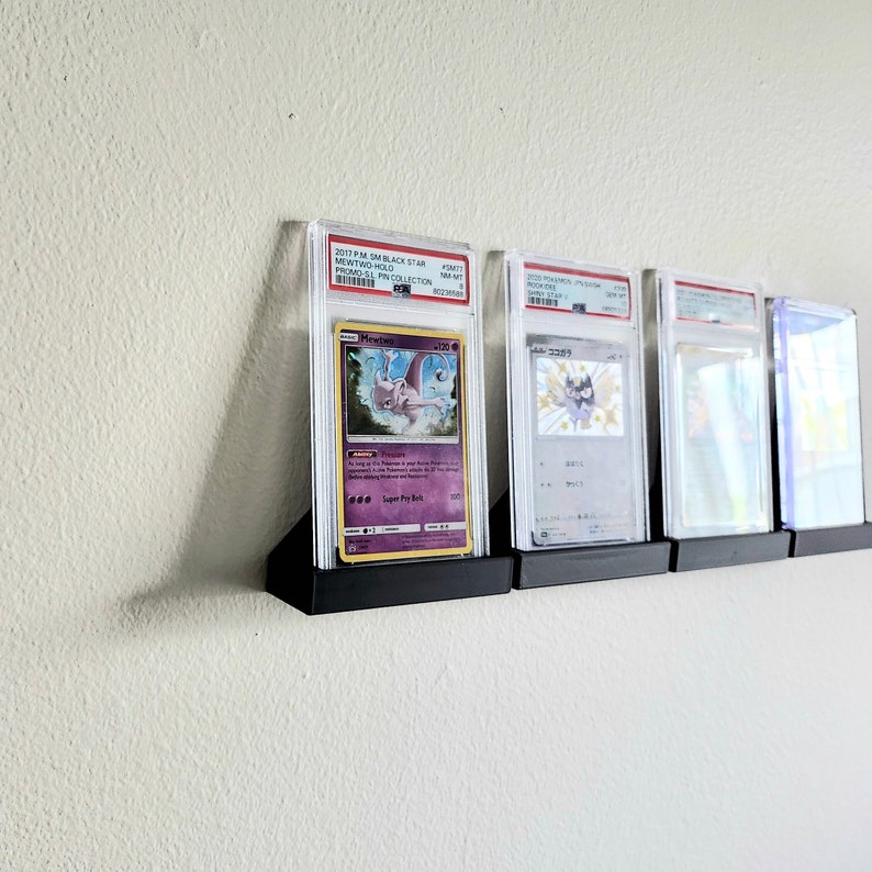 Puede incluir: Cuatro cartas de Pok&eacute;mon clasificadas se muestran en un soporte de pared negro. Las cartas est&aacute;n encerradas en soportes de pl&aacute;stico transparente con una etiqueta blanca en la parte superior. Las cartas son todas diferentes, pero todas presentan personajes de Pok&eacute;mon. Las cartas est&aacute;n etiquetadas con el texto "2017 PSA 10 BLACK STAR MEWTWO GOLD STAR 1ST EDITION PSA 10 COLLECTION", "2020 PSA 10 JAPANESE CHARIZARD VMAX PSA 10 COLLECTION", "2020 PSA 10 JAPANESE CHARIZARD VMAX PSA 10 COLLECTION" y "2020 PSA 10 JAPANESE CHARIZARD VMAX PSA 10 COLLECTION".
