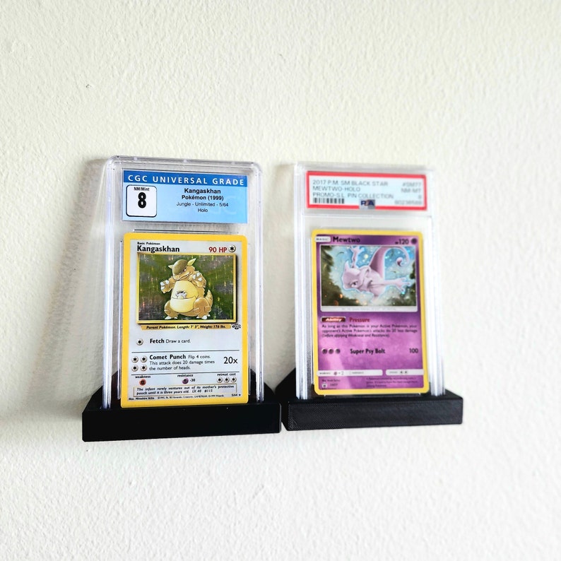 Puede incluir: Dos cartas de Pok&eacute;mon clasificadas en estuches protectores. La carta de la izquierda es un Kangaskhan del set Jungle con una calificaci&oacute;n CGC de 8. La carta de la derecha es un Mewtwo de la colecci&oacute;n promocional El Pin de Pokemon Black Star 2017.