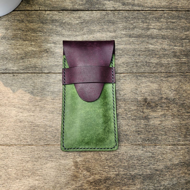 May include: A handmade leather phone case in shades of green and purple. The case features a flap closure and a strap detail. The edges are stitched with black thread. The case is shown on a wooden surface.