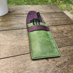 May include: A handcrafted leather pen case in shades of green and purple. The case is rectangular with a rounded top and features a dark purple flap and a green body. The case holds several black pens.