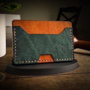 May include: A handcrafted leather card wallet in a bi-color design. The top section is a warm orange, while the lower section is a deep teal. The wallet features a stitched border and a debossed wolf emblem. It is displayed on a black stand.
