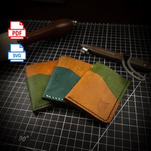 Può includere: Tre portafogli in pelle fatti a mano nei toni del verde, verde acqua e marrone, con tasche a colori contrastanti. Un portafoglio presenta un disegno a testa di lupo. I portafogli sono su una superficie a griglia con strumenti per la lavorazione della pelle.