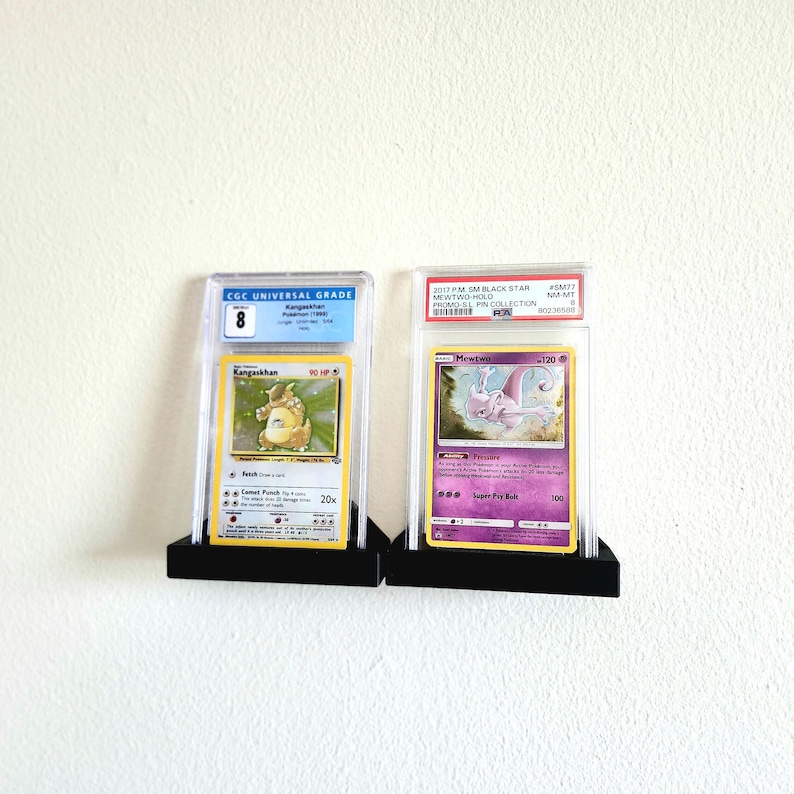 Puede incluir: Dos cartas de Pok&eacute;mon clasificadas en estuches protectores. La carta de la izquierda es una carta de Kangaskhan clasificada con un 8 por CGC. La carta de la derecha es una carta de Mewtwo clasificada con un 8 por PSA. Ambas cartas est&aacute;n en estuches negros con un fondo negro.
