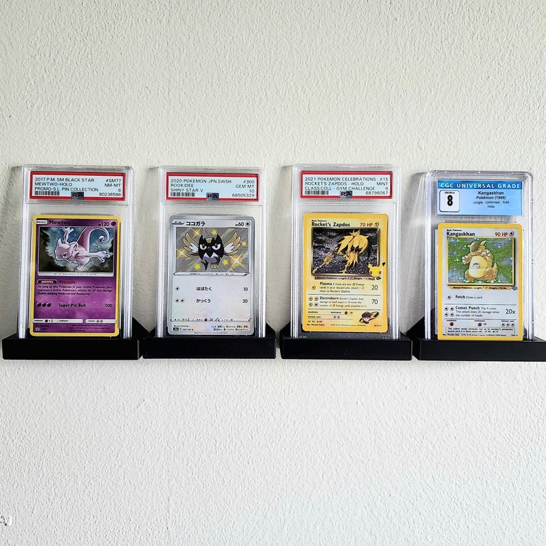 Puede incluir: Cuatro cartas de intercambio de Pok&eacute;mon se muestran en estuches protectores en una pared blanca. Las cartas est&aacute;n calificadas y presentan diferentes personajes de Pok&eacute;mon, incluidos Mewtwo, Rookidee, Rocket's Zapdos y Kangaskhan.