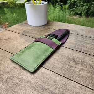 May include: A handmade leather pen case in green and purple, with black pens inside. The case is rectangular with a flap closure and stitched edges. The case is sitting on a wooden surface.