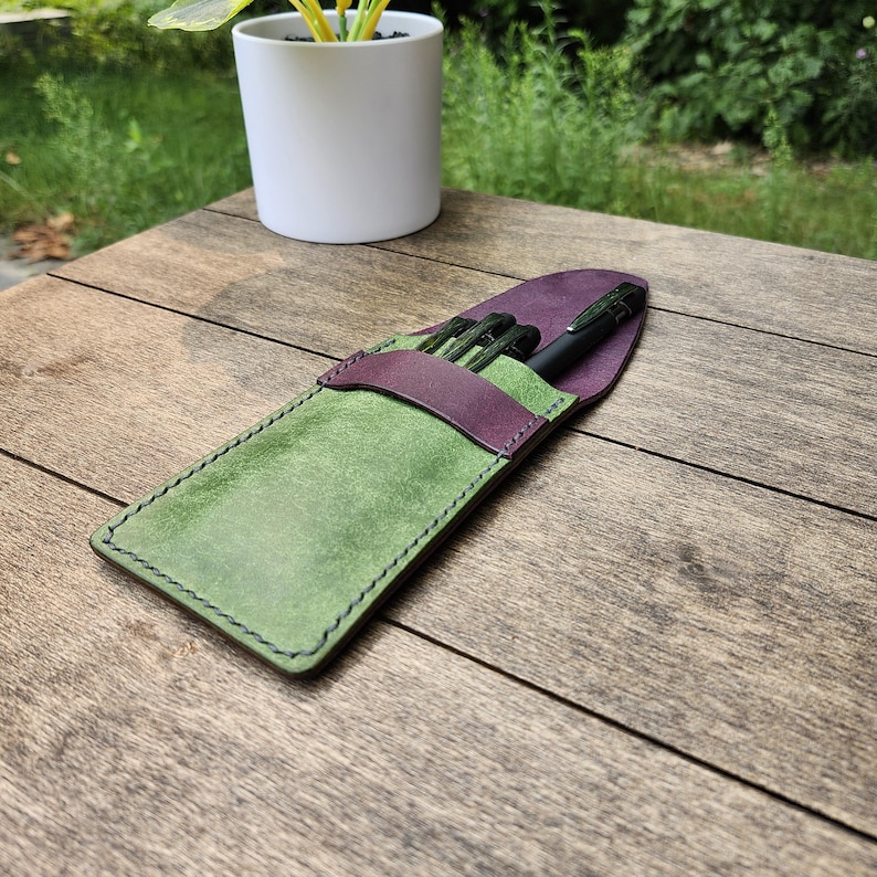 May include: A handmade leather pen case in green and purple, with black pens inside. The case is rectangular with a flap closure and stitched edges. The case is sitting on a wooden surface.