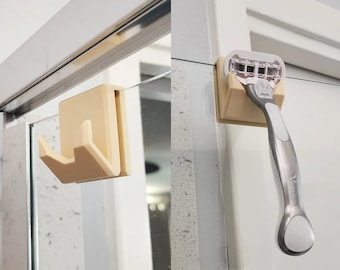 May include: Two beige plastic hooks for hanging items in a bathroom. One hook is designed to hold a razor and the other is a simple hook.