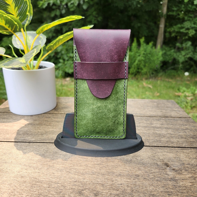 May include: A handcrafted leather card holder in shades of green and purple. The front is a vibrant green, while the flap and strap are a deep purple. The edges are stitched with a contrasting thread. The holder is displayed on a black stand.