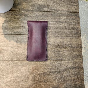 May include: A deep burgundy leather glasses case with a stitched border. The case is rectangular with a folded top. The leather has a smooth texture and a rich, dark color. The case is designed to protect eyeglasses.