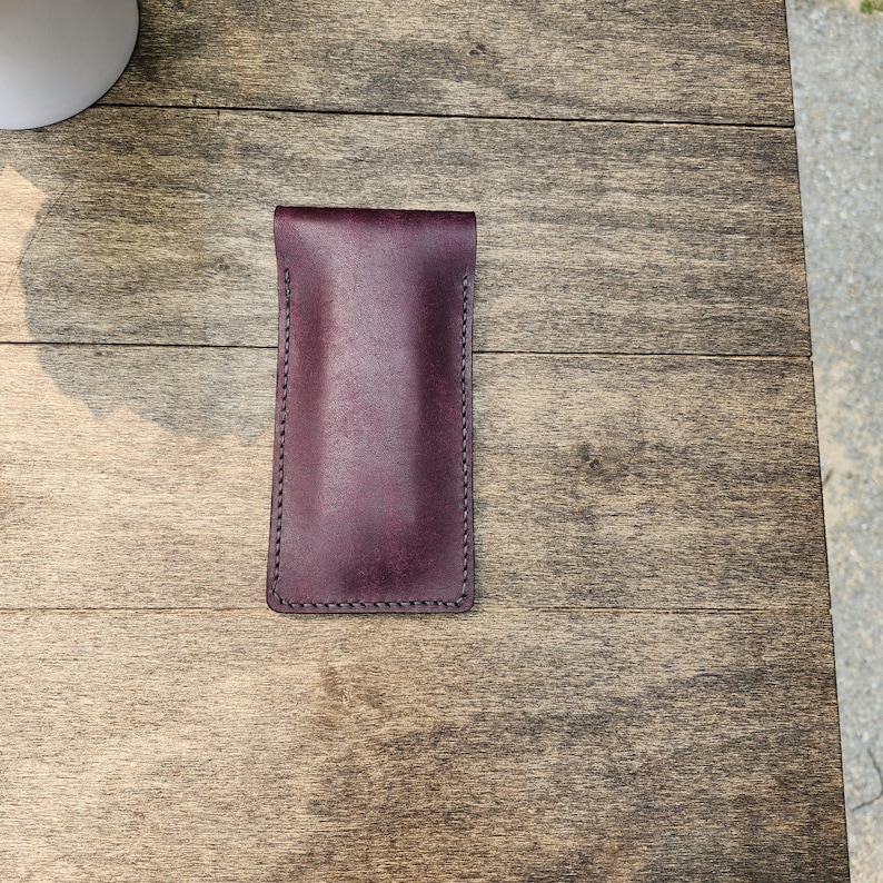 May include: A deep burgundy leather glasses case with a stitched border. The case is rectangular with a folded top. The leather has a smooth texture and a rich, dark color. The case is designed to protect eyeglasses.