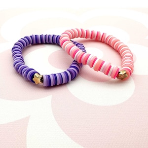 Könnte beinhalten: Zwei bunte Armbänder aus übereinander gestapelten, runden Perlen. Ein Armband ist rosa, weiß und hellrosa, das andere ist lila, lavendel und helllila. Beide Armbänder haben einen goldenen Sternanhänger.