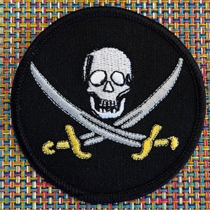 May include: Black embroidered patch with a white skull and crossbones. The skull has two eye sockets and a toothy grin. The crossbones are silver with yellow handles.