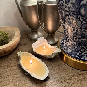 May include: Two oyster shell candles with a single wick each, burning on a wooden surface. The candles are surrounded by silver vases and a blue and white patterned lamp.