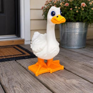 May include: A white goose figurine with a long neck, textured feathers, and bright orange webbed feet. The goose has a prominent orange beak and large blue eyes. The figure is constructed from a smooth, molded material and stands upright on a flat, rectangular orange base.