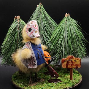 May include: A taxidermied duck wearing a hockey mask and a blue and white flannel shirt, holding a chainsaw. The duck is standing on a green base with a sign that reads "Camp Blood" in red letters.
