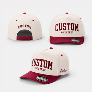 Könnte beinhalten: Cremefarbene Baseballkappe mit weinrotem Schirm und Akzenten. Das Wort "CUSTOM" ist weinrot auf der Vorder- und Rückseite gestickt. Der Text "YOUR TEXT" befindet sich unter dem Wort "CUSTOM" auf der Vorderseite. Die Kappe hat einen Snapback-Verschluss.