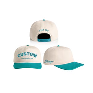 May include: A beige and teal baseball cap with the text "CUSTOM Los Angeles, Ca" on the front and "Savage" on the side. The back of the cap has the text "UTAH 2025".