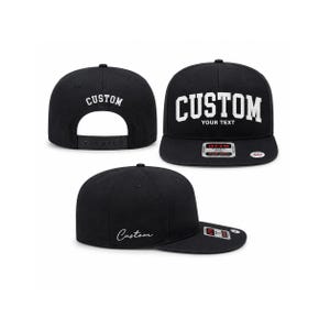 May include: Black flat-brimmed baseball cap with the word "CUSTOM" in white block letters. The cap is shown from multiple angles, including the front, back, and side. The text "YOUR TEXT" is below the word "CUSTOM" on the front.