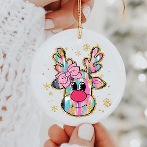 May include: A round, white Christmas ornament featuring a colorful reindeer design. The reindeer has a pink nose, a pink bow, and gold glitter accents. The ornament is held by a hand and has a gold string for hanging.