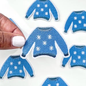 May include: Six blue embroidered iron-on patches shaped like sweaters with white snowflake designs.