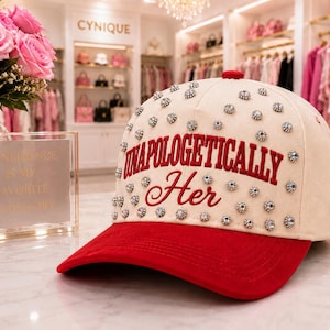 May include: A cream-colored baseball cap with a red brim, featuring the red embroidered text "UNAPOLOGETICALLY Her" on the front. The cap is decorated with numerous small, round, silver-toned studs with clear crystals scattered across the crown.