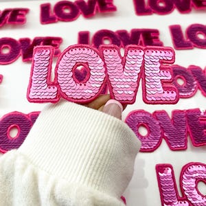 May include: Pink sequined iron-on patch that says "LOVE" in all capital letters. The patch is being held by a hand wearing a white sweater.