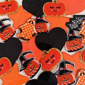 May include: A collection of embroidered and sequined patches featuring orange pumpkins with smiling faces, black hearts, orange stars, white ghosts with orange polka dots, and pumpkins wearing black witch hats with silver buckles.
