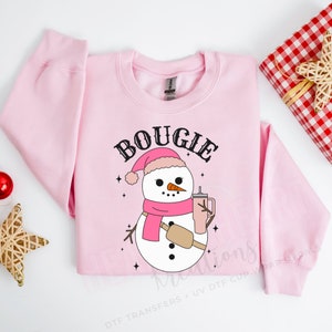 May include: Pink sweatshirt with a snowman graphic and the text "BOGIE" in black. The snowman is wearing a pink hat and scarf and holding a pink cup.