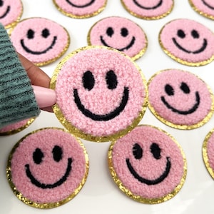 May include: Pink smiley face patches with black embroidered details and a gold glitter border. The patches have a fuzzy texture and are round in shape. Several patches are visible.
