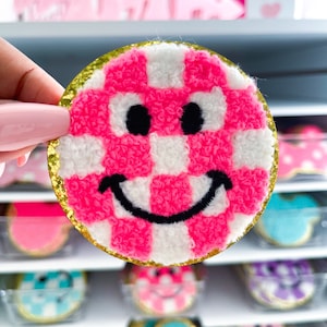May include: A round, pink and white checkered smiley face patch with a gold glitter border. The smiley face has black eyes and a curved black mouth. Other smiley face cookies are visible in the background.